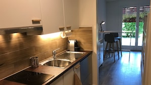 A modern kitchen with a stainless steel sink, a built-in stove, and a wooden backsplash. There is a small island with a stool and a bar area visible through a glass door. - Large Studio Near Epfl And Lausanne City Center (Ecublens)
