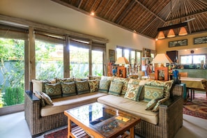 A spacious living area with a large sofa, a glass coffee table, and a dining table. There are large windows providing natural light and a view of greenery outside.