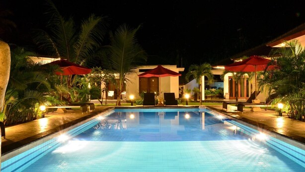 A nighttime pool area with a rectangular pool, red umbrellas, and palm trees. - Explore Lombok From Dream Estate (Senggigi)