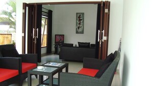 A living room with wicker furniture, a glass coffee table, and a black sofa with red cushions. - Explore Lombok From Dream Estate (Senggigi)