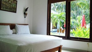 A bedroom with a bed, a wall-mounted lamp, and a large window with a view of palm trees and outdoor furniture. - Explore Lombok From Dream Estate (Senggigi)