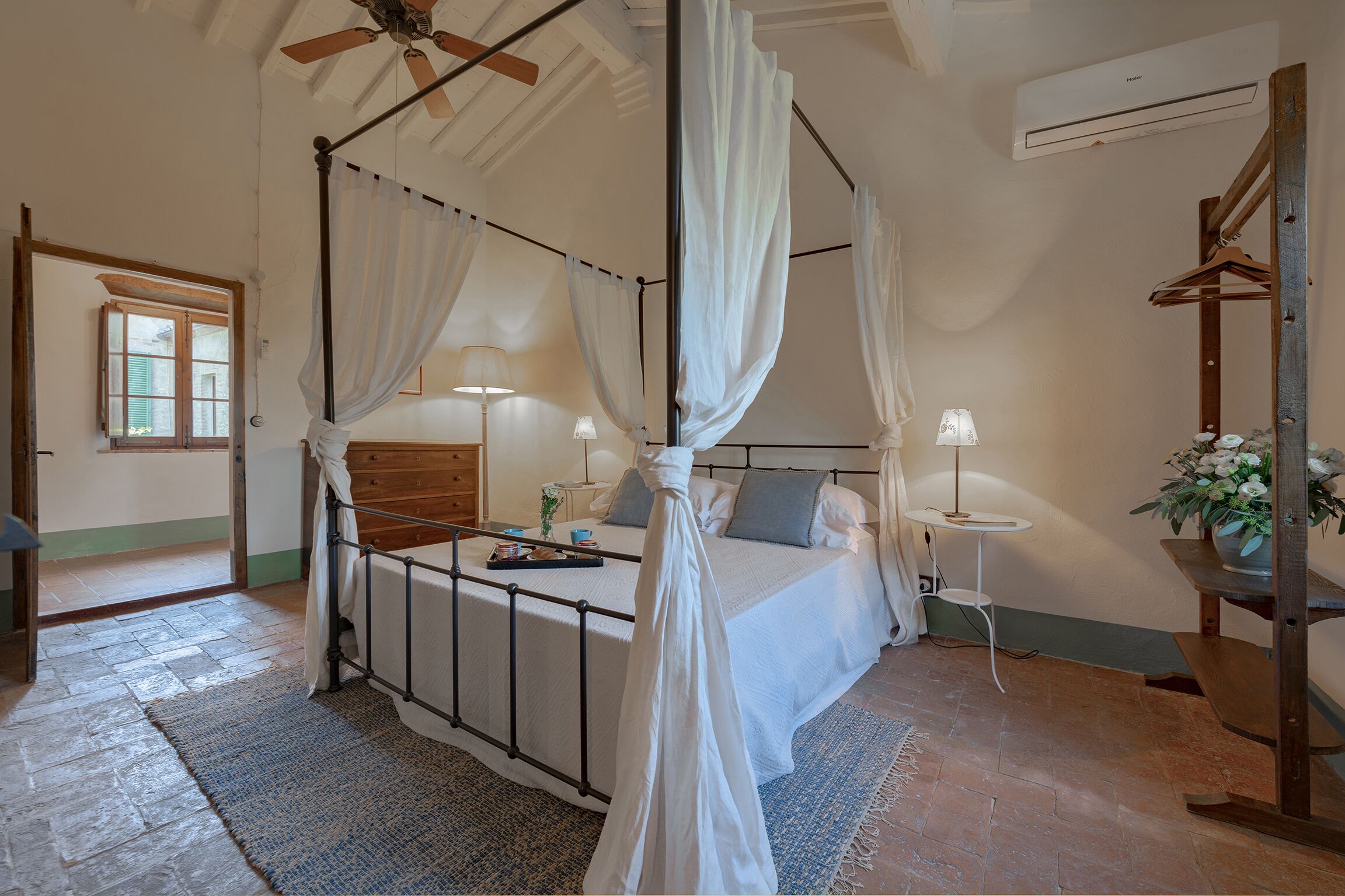 A bedroom with a four-poster bed, a wooden wardrobe, a bedside table with a lamp, and a small potted plant.