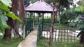 Un pont en bois, abrité, mène à un petit pavillon entouré d’une végétation luxuriante et bordé par un plan d’eau paisible.