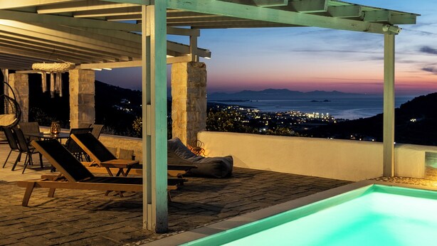 Zona de piscina exterior con sillas de descanso y una pérgola, con vistas a un paisaje urbano al atardecer.