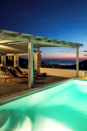 Zona de piscina exterior con sillas de descanso y una pérgola, con vistas a un paisaje urbano al atardecer.