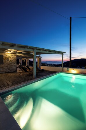 A swimming pool with a stone wall and lounge chairs under a pergola, overlooking a scenic view.