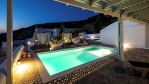 A swimming pool with a stone border, under a pergola, surrounded by plants and lights. - Villa Thetis With Private Pool (Paros)