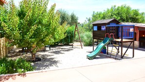 Ein Spielplatz mit Rutsche und Schaukeln, umgeben von Bäumen und einem Gebäude mit blauem Dach.