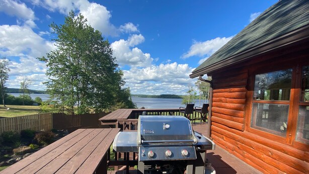 Un terrazzo in legno con griglia, tavolo da picnic e sedie, con vista su un lago.