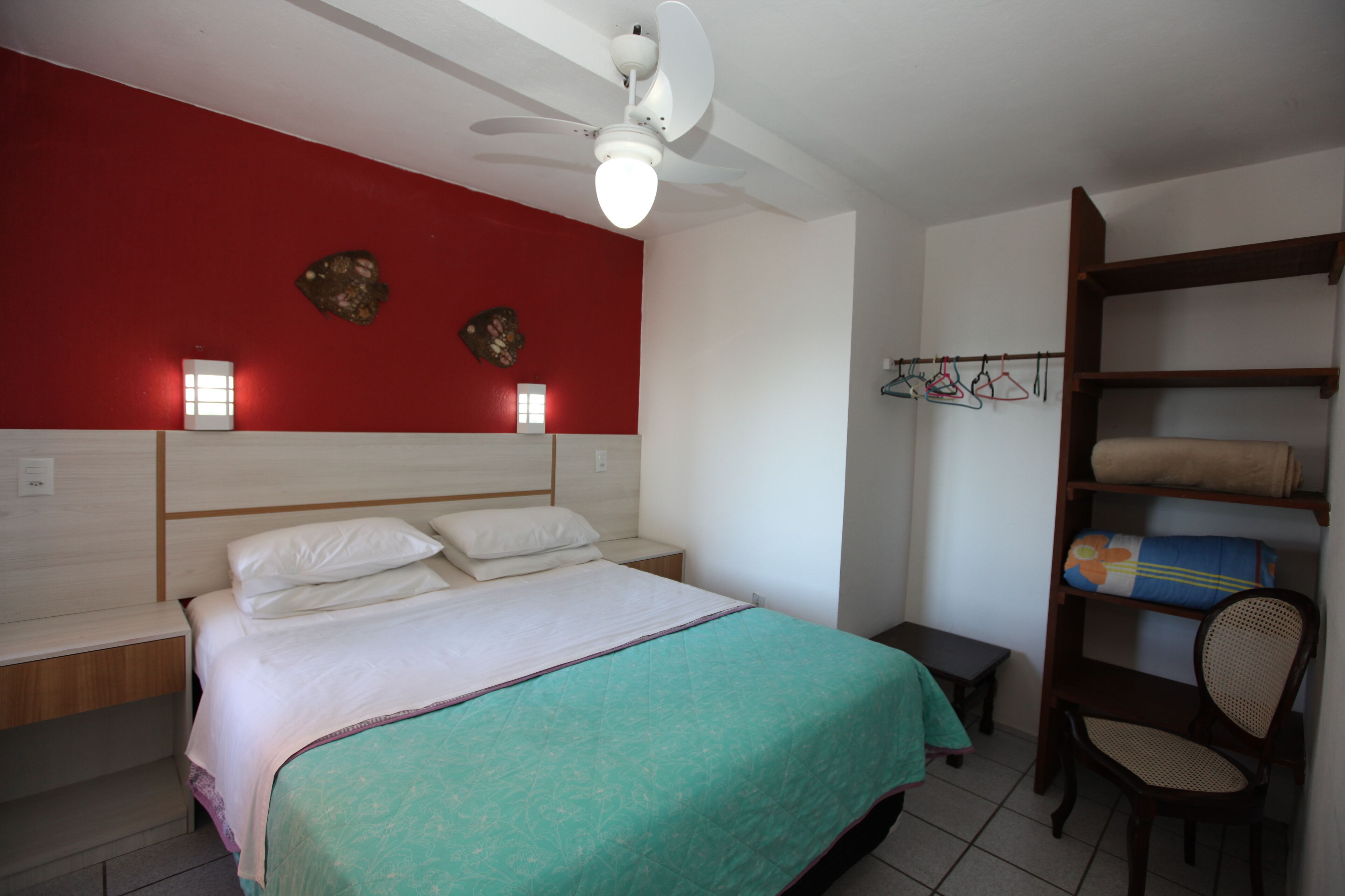A bedroom with a bed, a nightstand, a chair, a shelf with towels, and a red wall with decorative items.