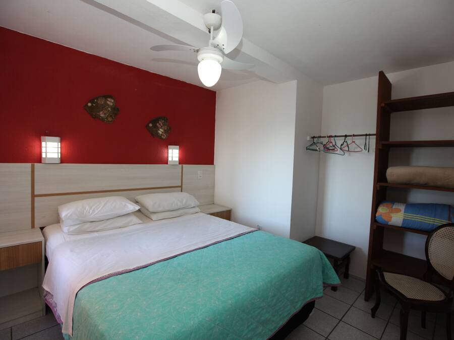 A bedroom with a bed, a nightstand, a chair, a shelf with towels, and a red wall with decorative items.