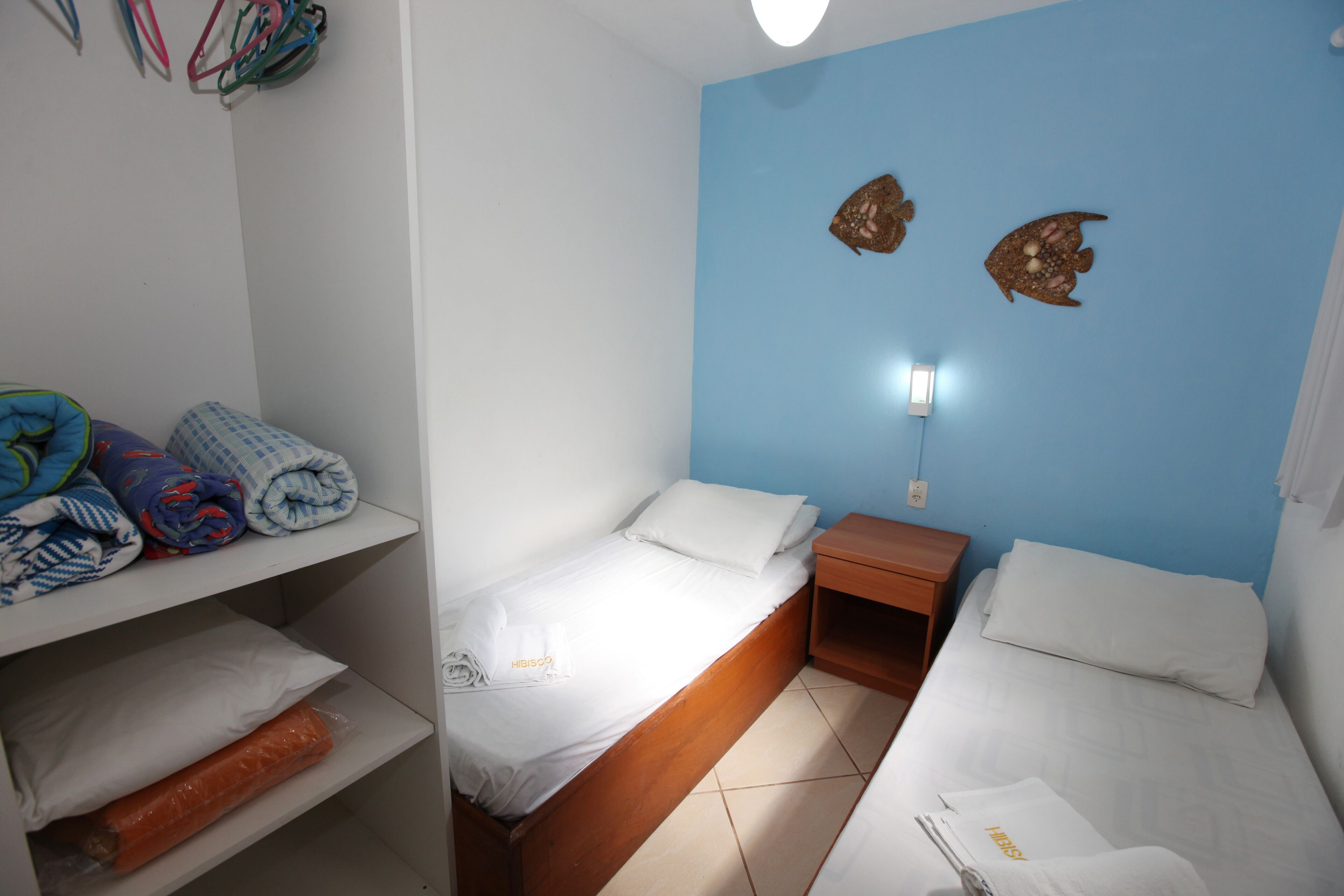 Una habitación pequeña y limpia con dos camas, una estantería con mantas dobladas y un muro decorado con dos adornos de peces.