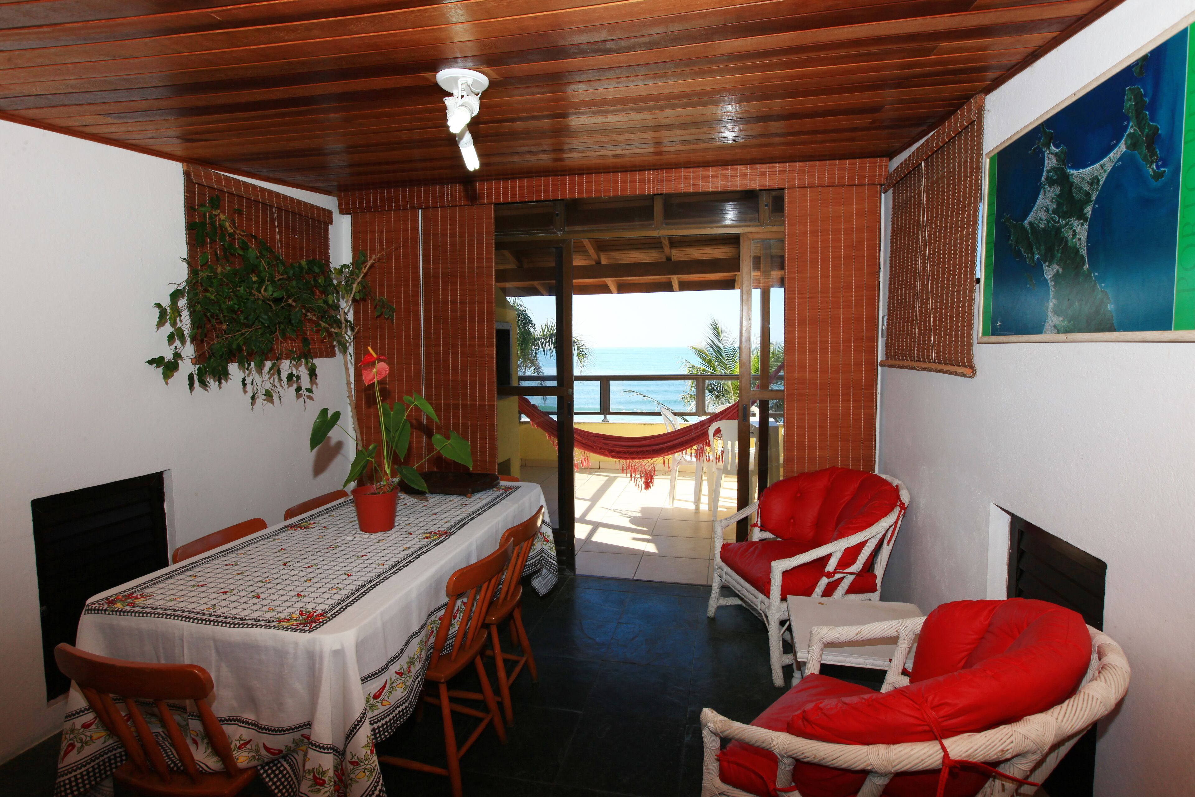 Uma área de refeições com uma mesa posta para quatro pessoas, rodeada por cadeiras de madeira, um vaso de planta e vista para o mar através de uma porta de vidro.