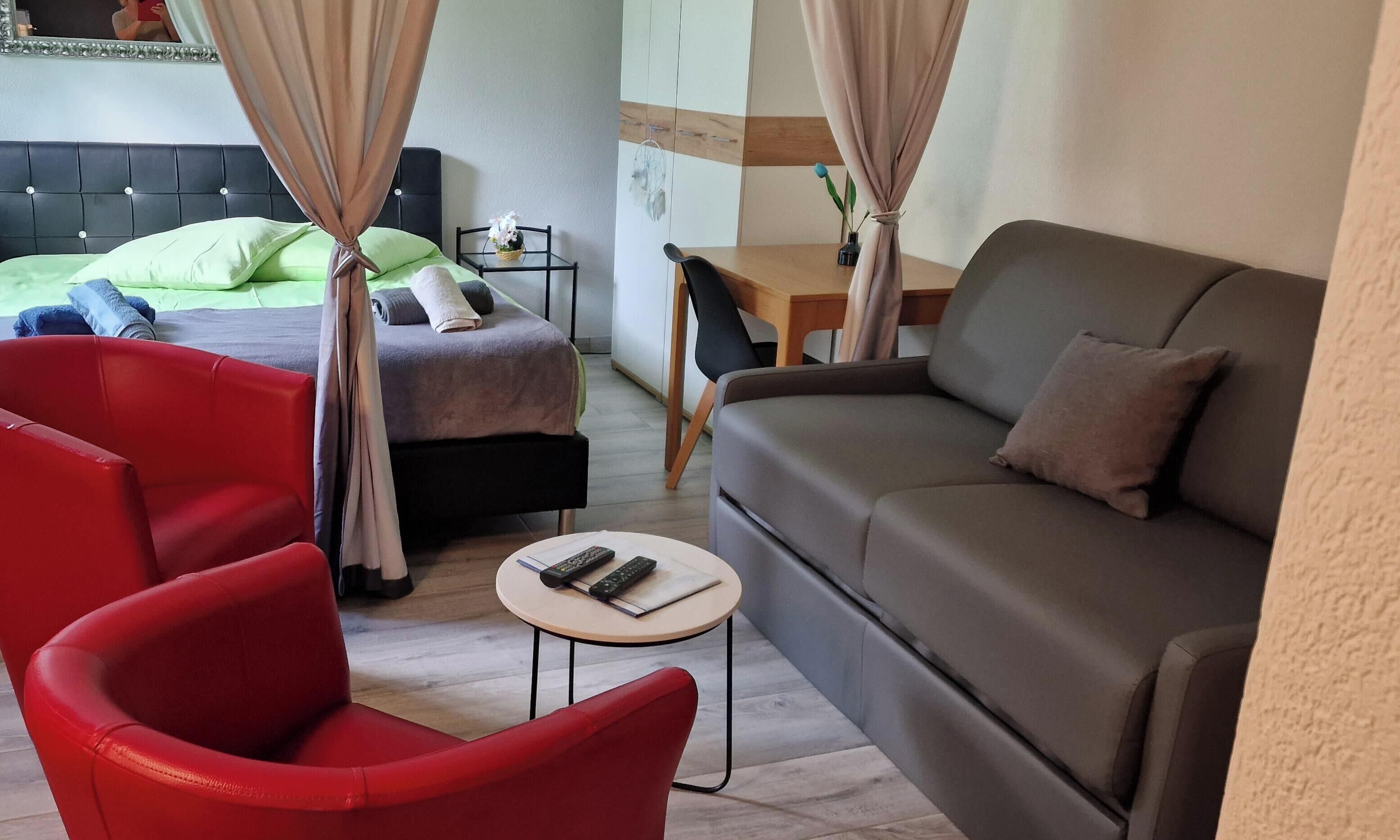 Ein Hotelzimmer mit einem Bett, einem roten Sessel, einem grauen Sofa, einem kleinen Tisch und einem Schreibtisch mit Stuhl.