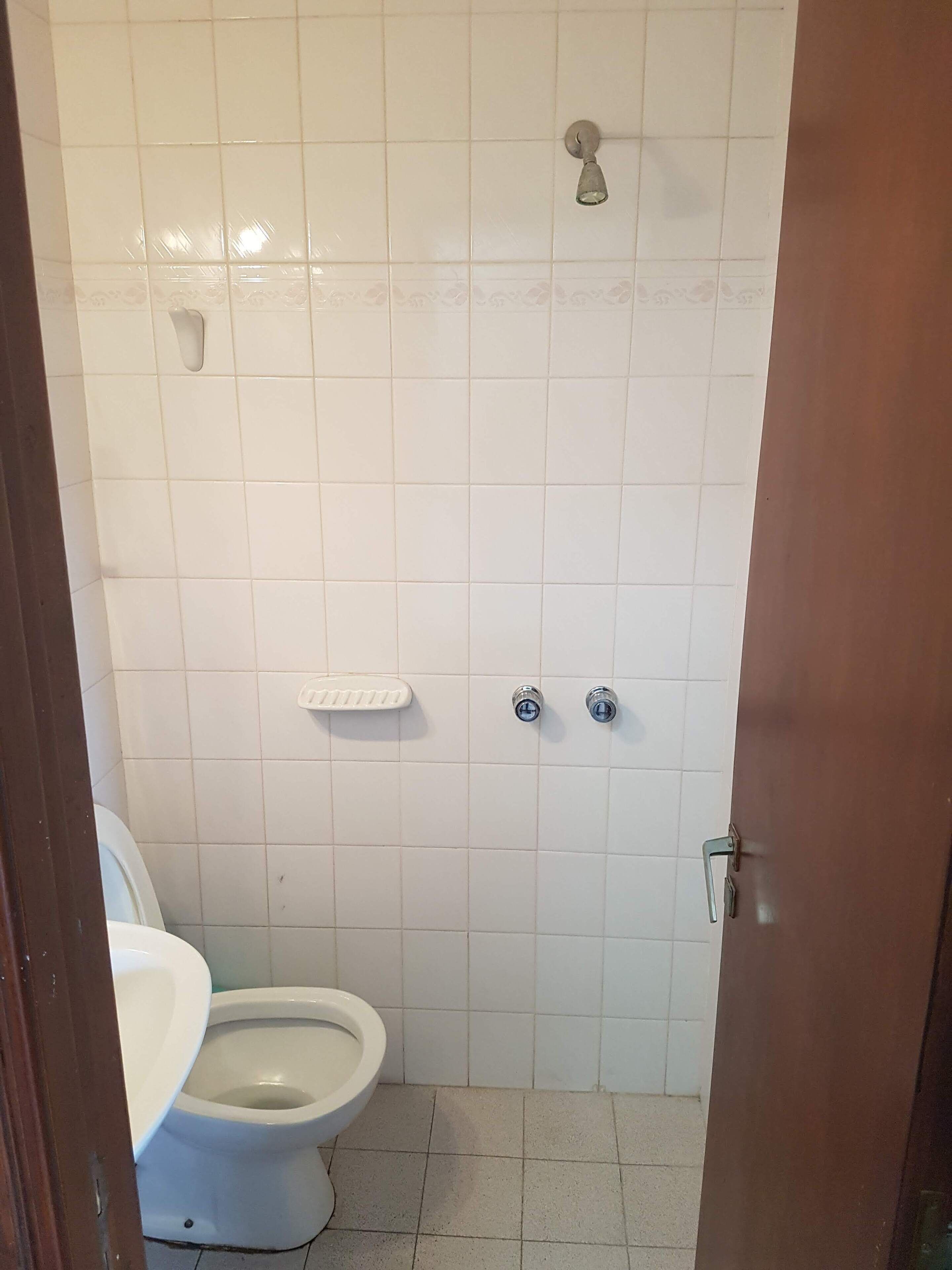 A small, clean bathroom with white tiles, a toilet, a shower, and a sink.