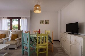Un comedor con una mesa dispuesta para dos personas, un televisor en un mueble y una ventana con cortinas.