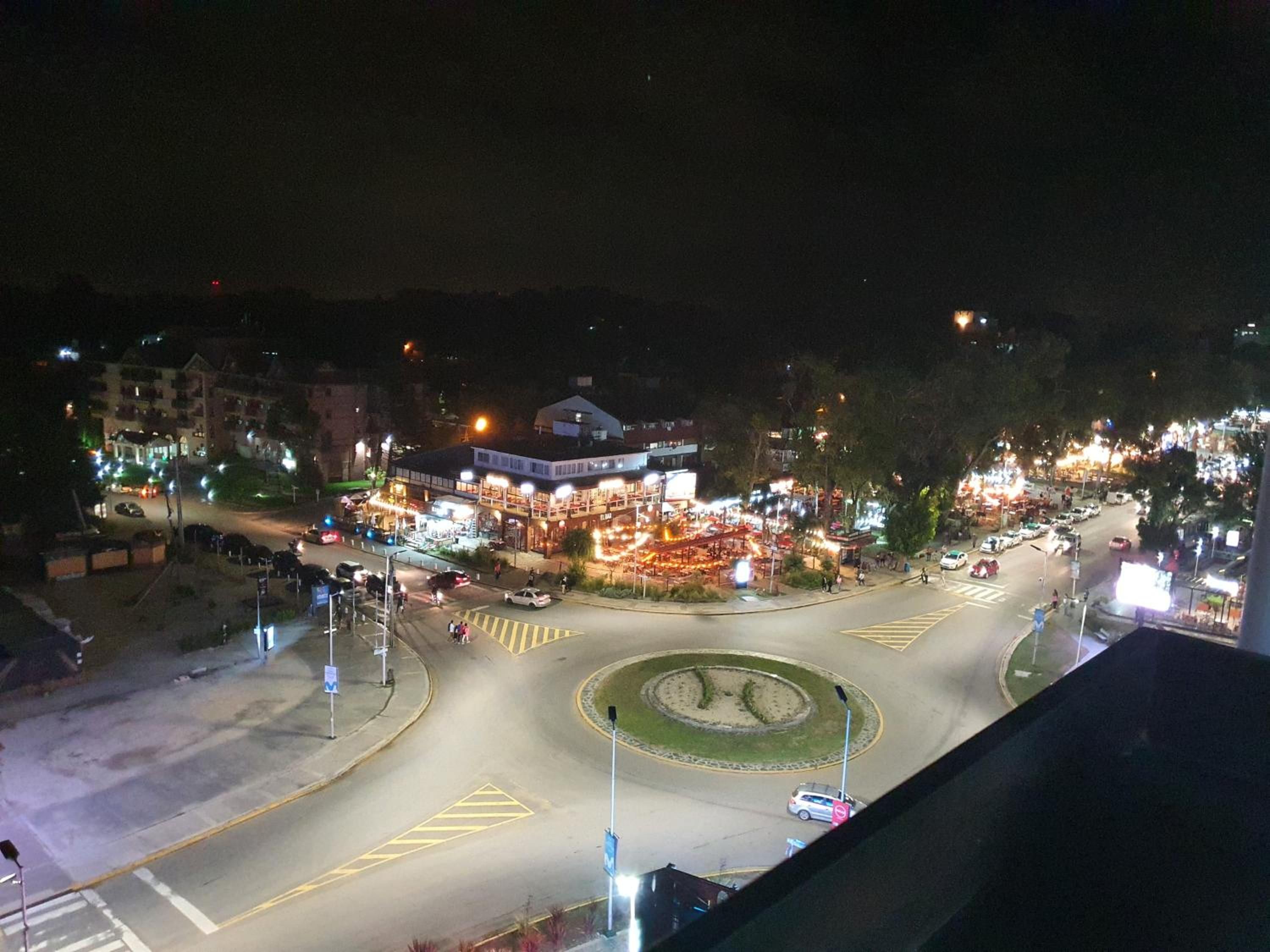 Vista noturna de um cruzamento circular com praça central, edifícios e postes de luz ao redor.