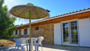 Een terras met een tafel en stoelen, een strooien parasol en een stenen gebouw met een pannendak en grote ramen.