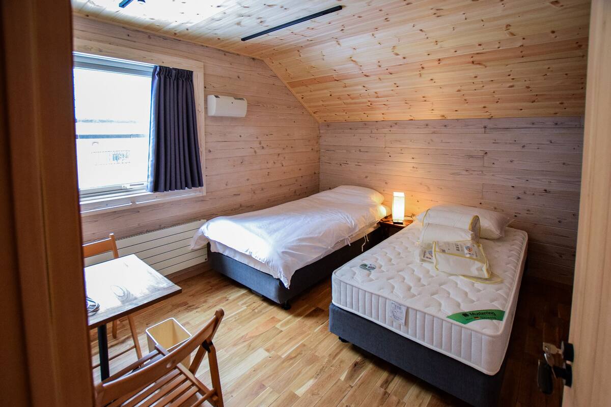 Una habitación pequeña y acogedora con paredes de madera y techo inclinado. Tiene una cama individual, una mesita de noche, una silla y una ventana con cortinas.