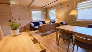 A cozy room with a wooden floor, a sofa, a dining table with chairs, a lamp, and a window with blinds. - Baan Hakuba (Hakuba)