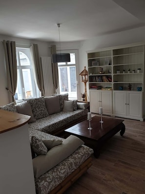 Un salon comprenant un canapé d’angle, une table basse, une bibliothèque et une fenêtre avec des rideaux.