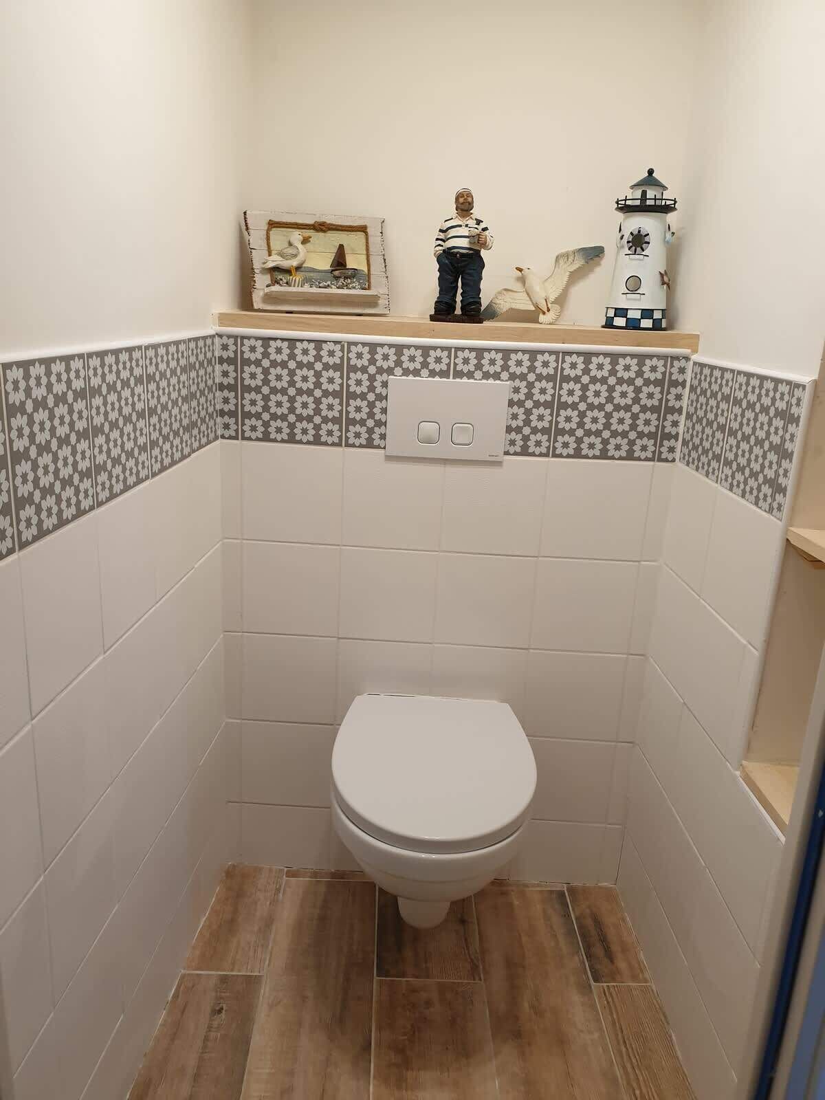 A small bathroom with a toilet, patterned tiles, a wooden shelf with decorative items, and a light switch.