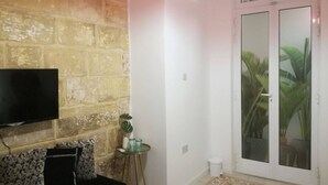 A living room with a TV mounted on a brick wall, a small round table with a plant, and a sliding glass door leading to a balcony with plants. - Private Room With Ensuite Groundfloor (Luqa)