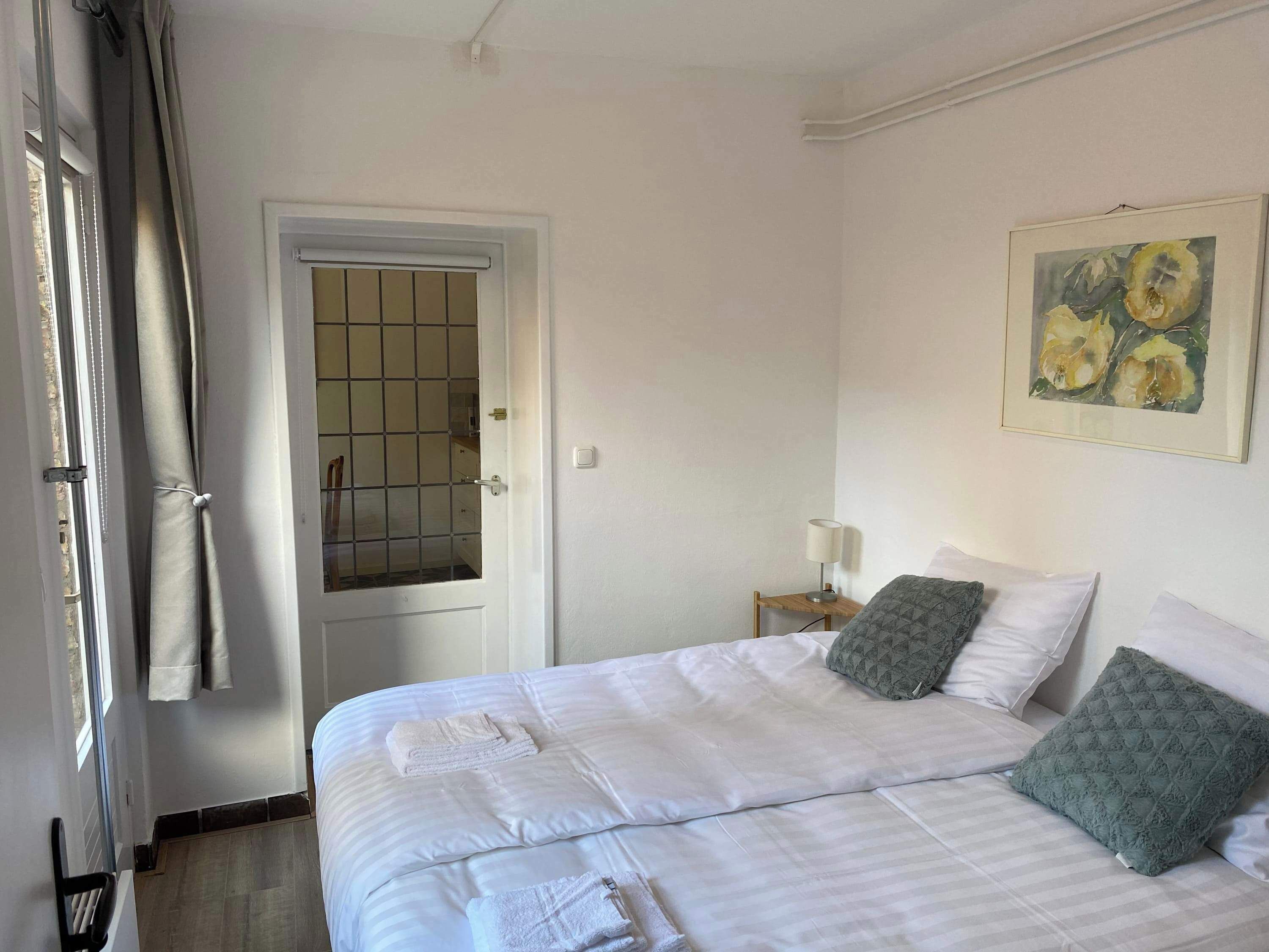 Une chambre d’hôtel avec un lit, une porte avec une fenêtre, une table de chevet avec une lampe et un tableau au mur.