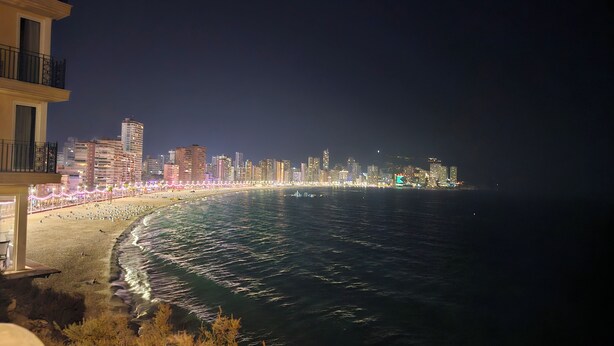 Une vue nocturne d’une ville côtière, avec des bâtiments illuminés et une plage.
