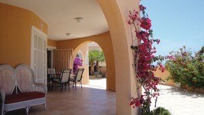 Une terrasse couverte avec une table et des chaises, un banc et une vue sur un jardin agrémenté de bougainvilliers.
