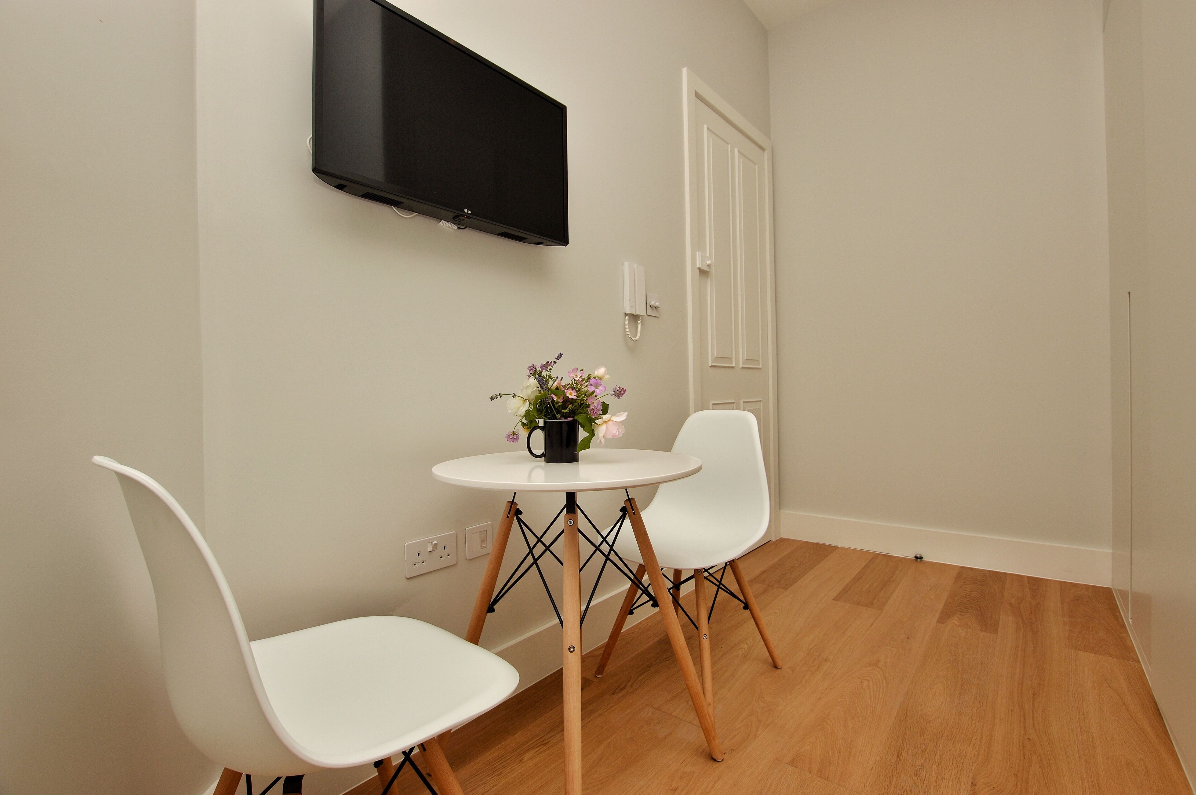 A small room with a white table and two white chairs, a flat-screen TV mounted on the wall, and a door with a white frame.