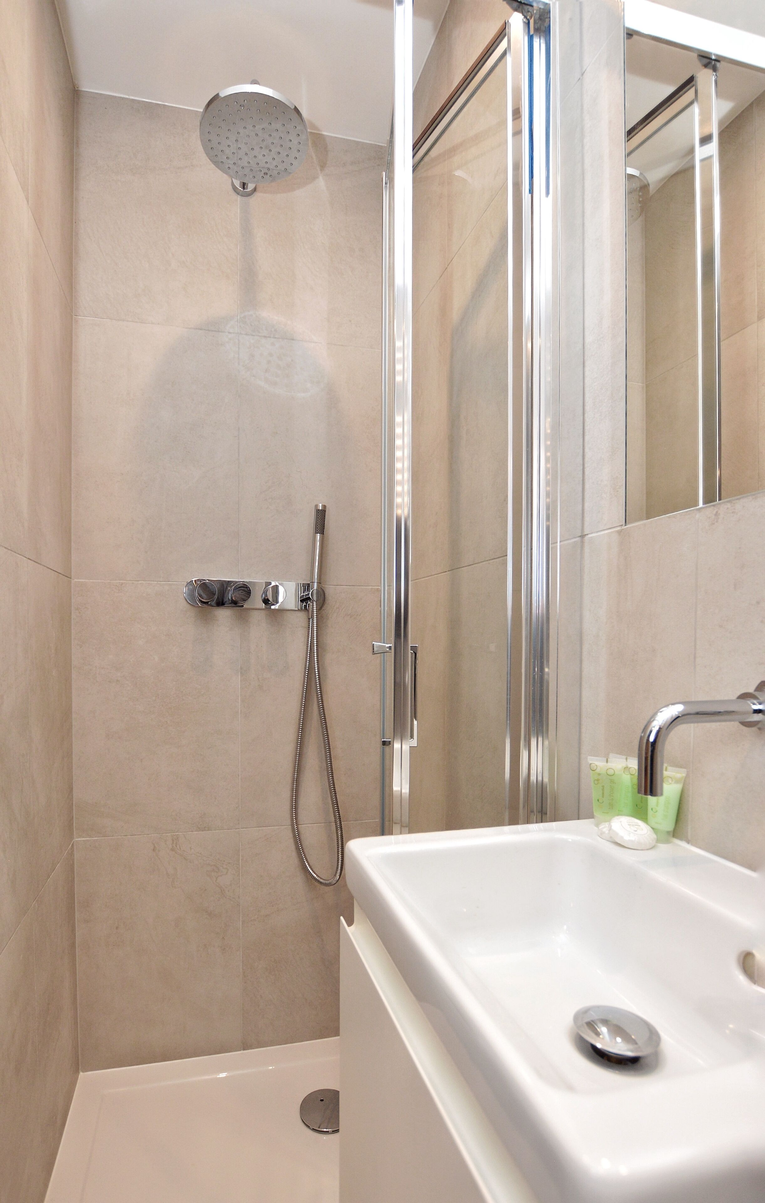 A modern shower with a handheld showerhead and a handheld hose, a white sink with a soap dispenser, and a mirror on the wall.