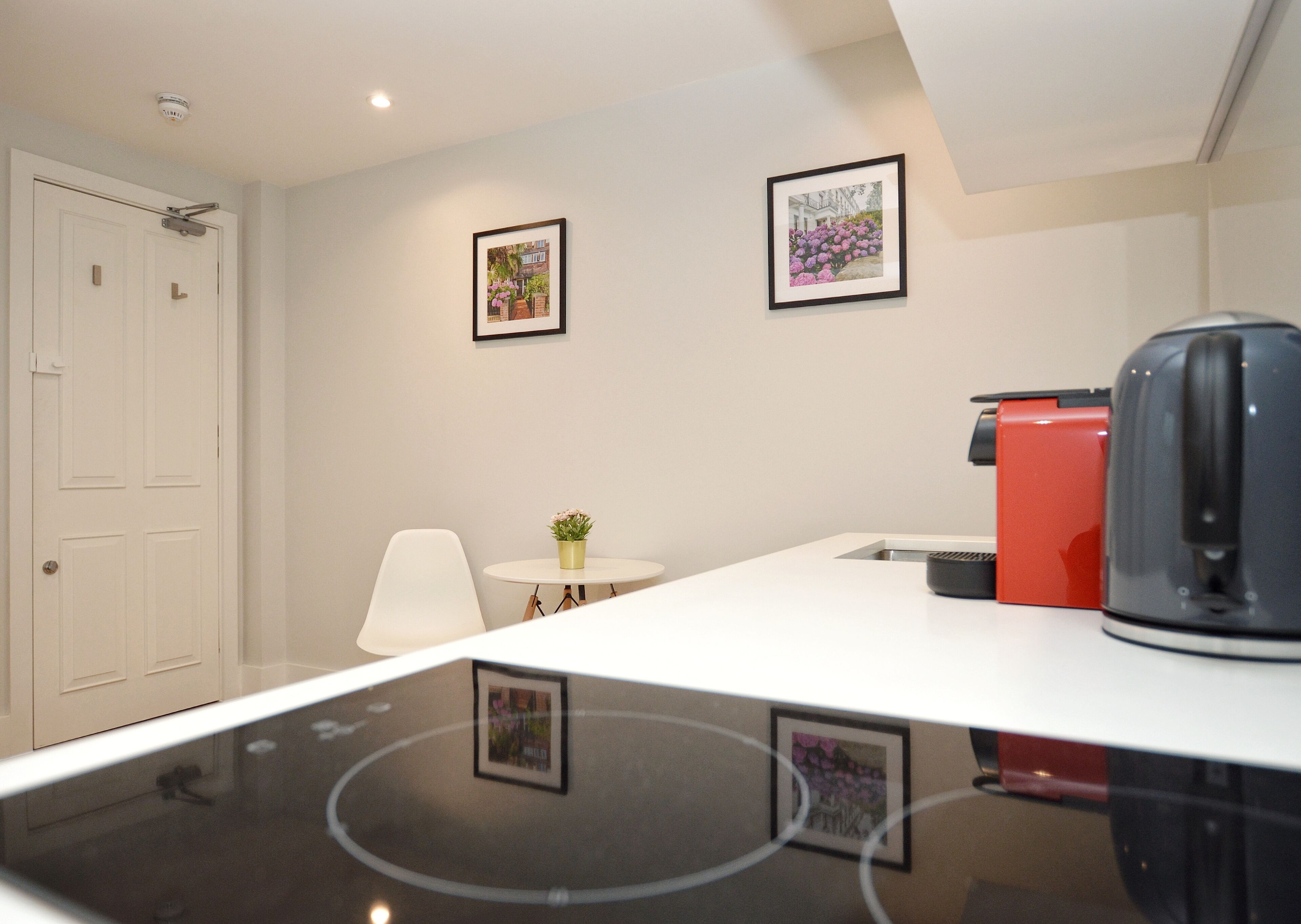 Une cuisine moderne avec un plan de travail blanc, une machine à café et un appareil électroménager rouge. On y trouve une petite table avec une plante en pot et un tableau encadré accroché au mur.