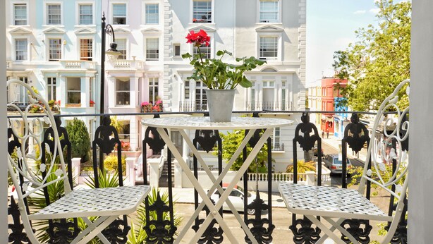 Ein Balkon mit weißen Metallstühlen und einem Tisch, ein Blumentopf mit roten Blüten und im Hintergrund mehrstöckige Gebäude.
