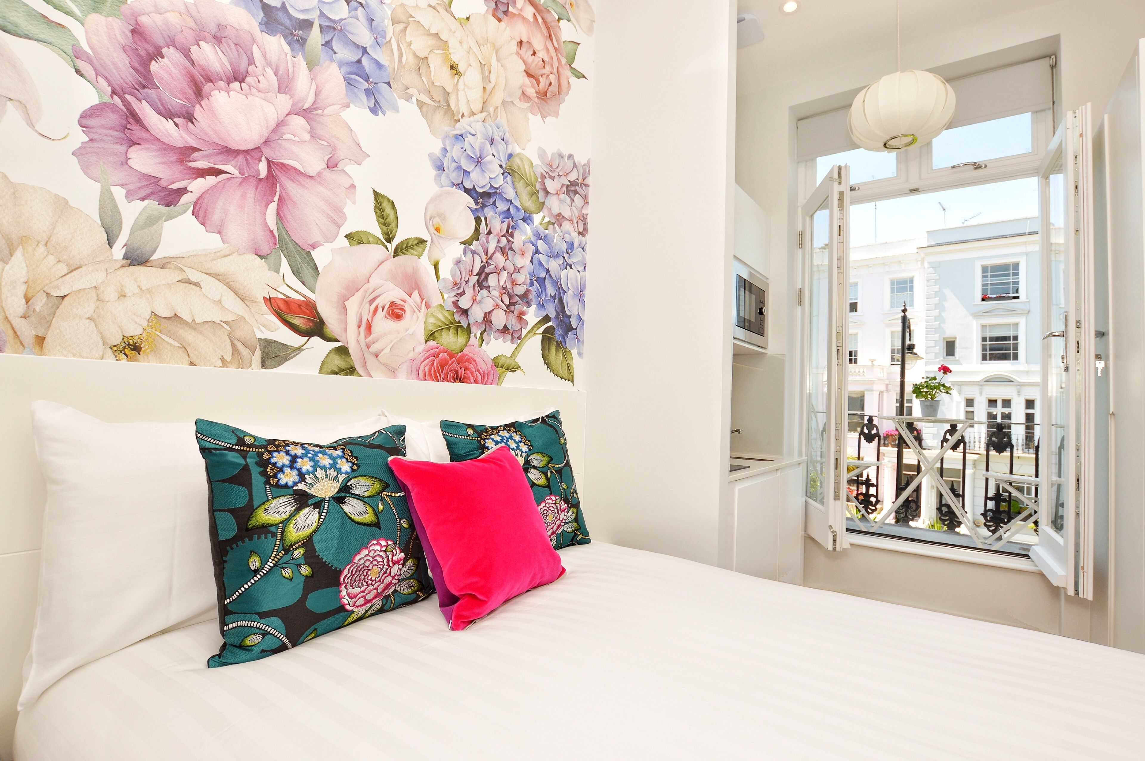 A bedroom with a large floral mural, a bed with white bedding, and a window with a view of buildings.