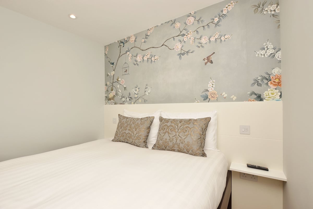 A hotel room with a bed, two pillows, a nightstand, a remote control, and a floral wall mural.