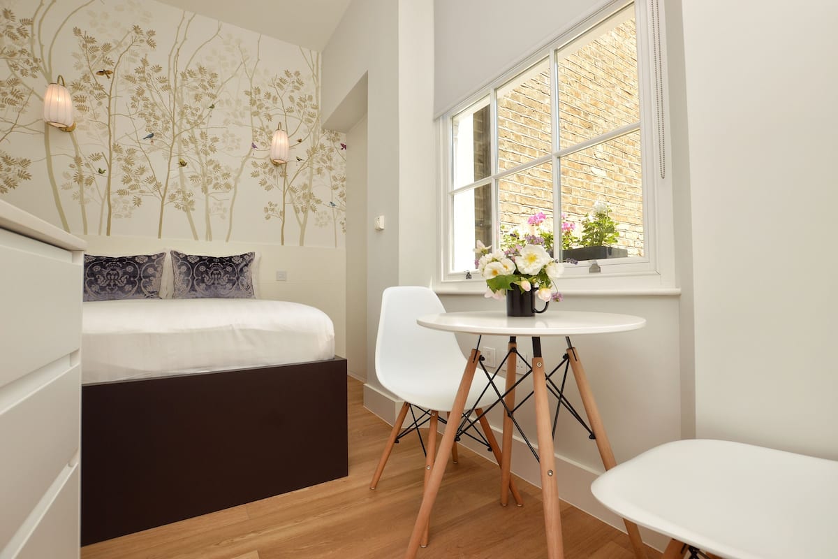 A modern room with a bed, white chairs, a small table with flowers, and a window with a view of a brick wall.