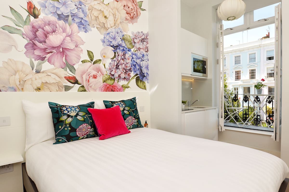 A bedroom with a large floral wallpaper, a white bed with two pillows, and a window with a view of buildings.