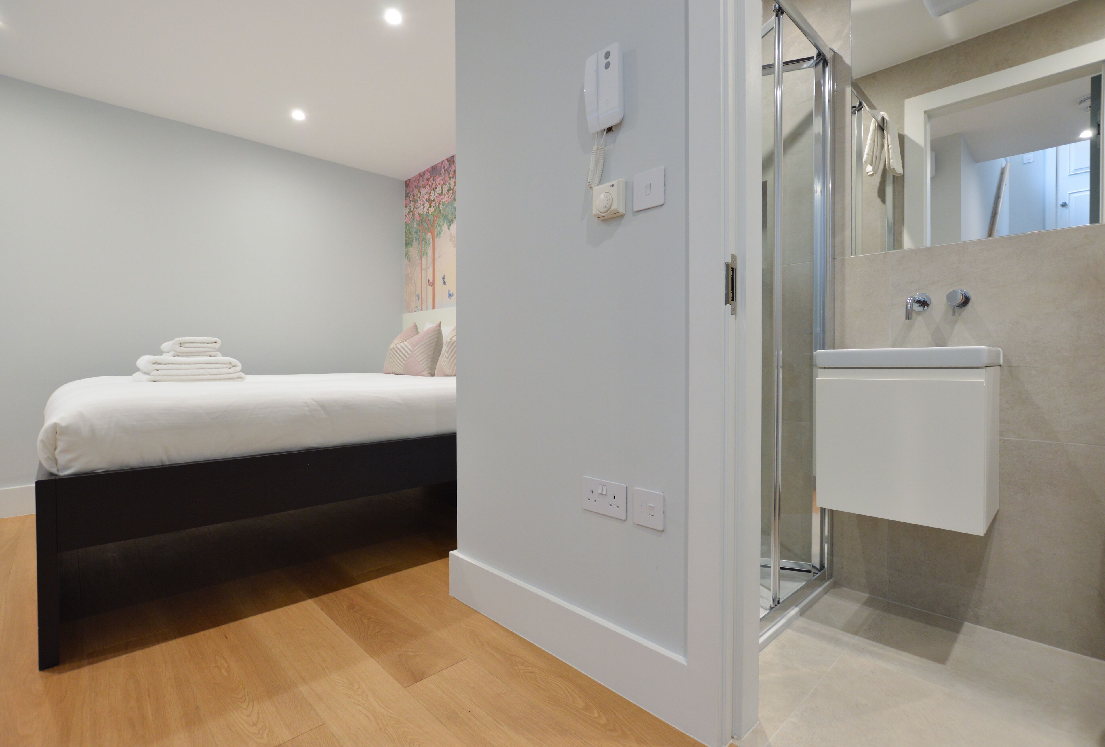 Een moderne slaapkamer met een bed, nachtkastje, douche en badkamer met wastafel en spiegel.