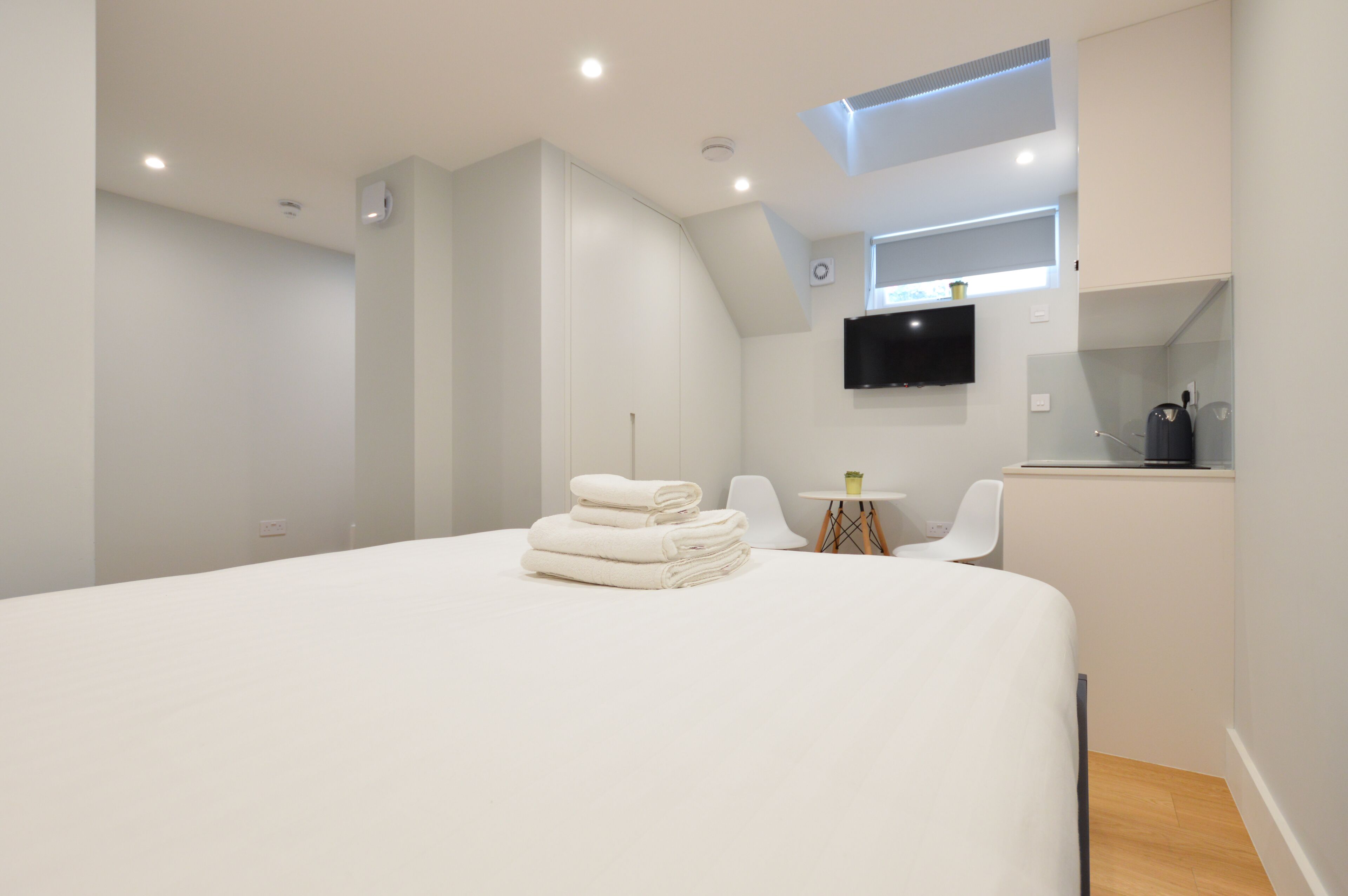 A modern hotel room with a large bed, a small table with chairs, a flat-screen TV mounted on the wall, and a kitchenette area.