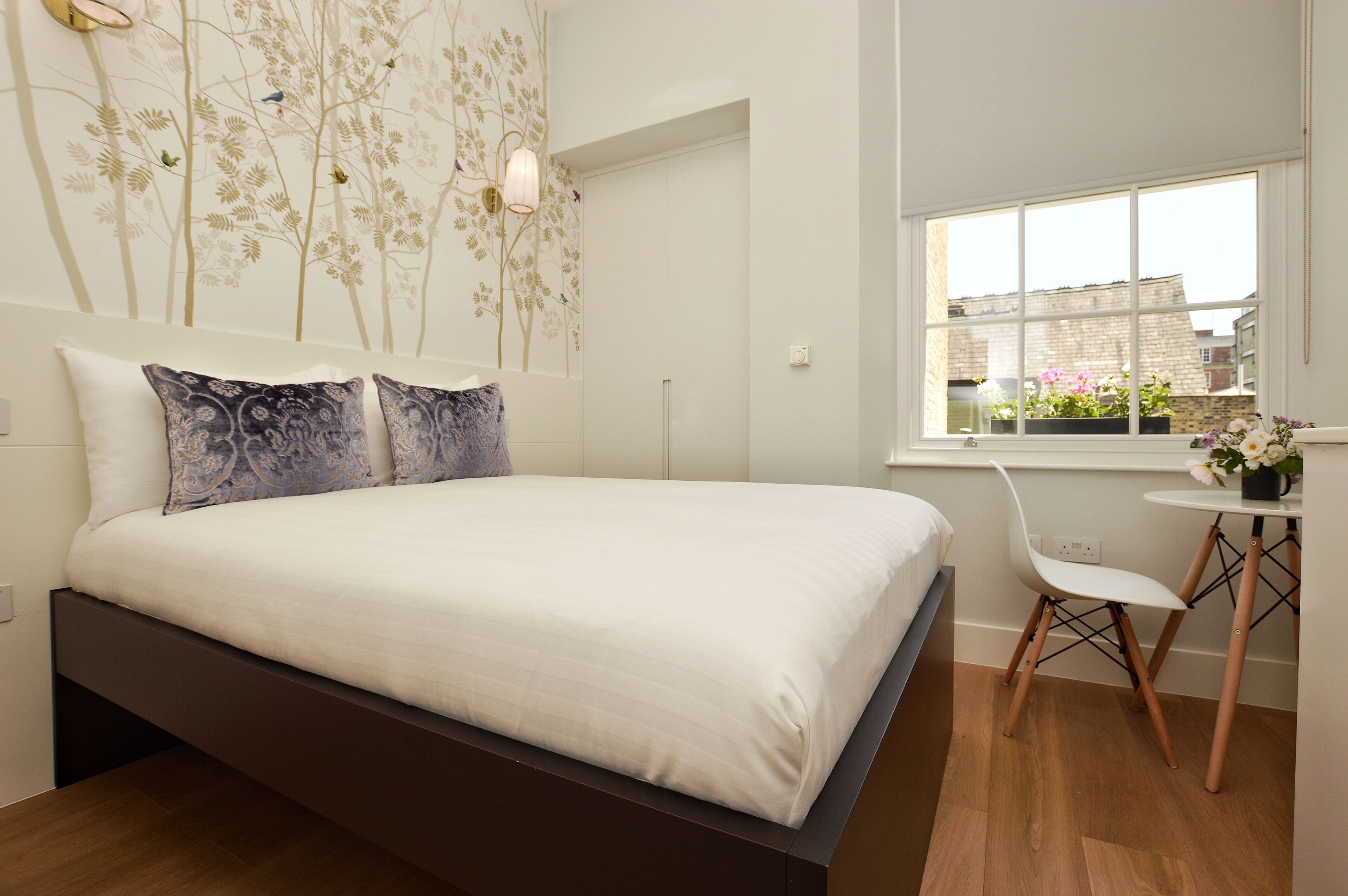 A bedroom with a bed, a window with a view of buildings, a chair, and a small table with a flower vase.