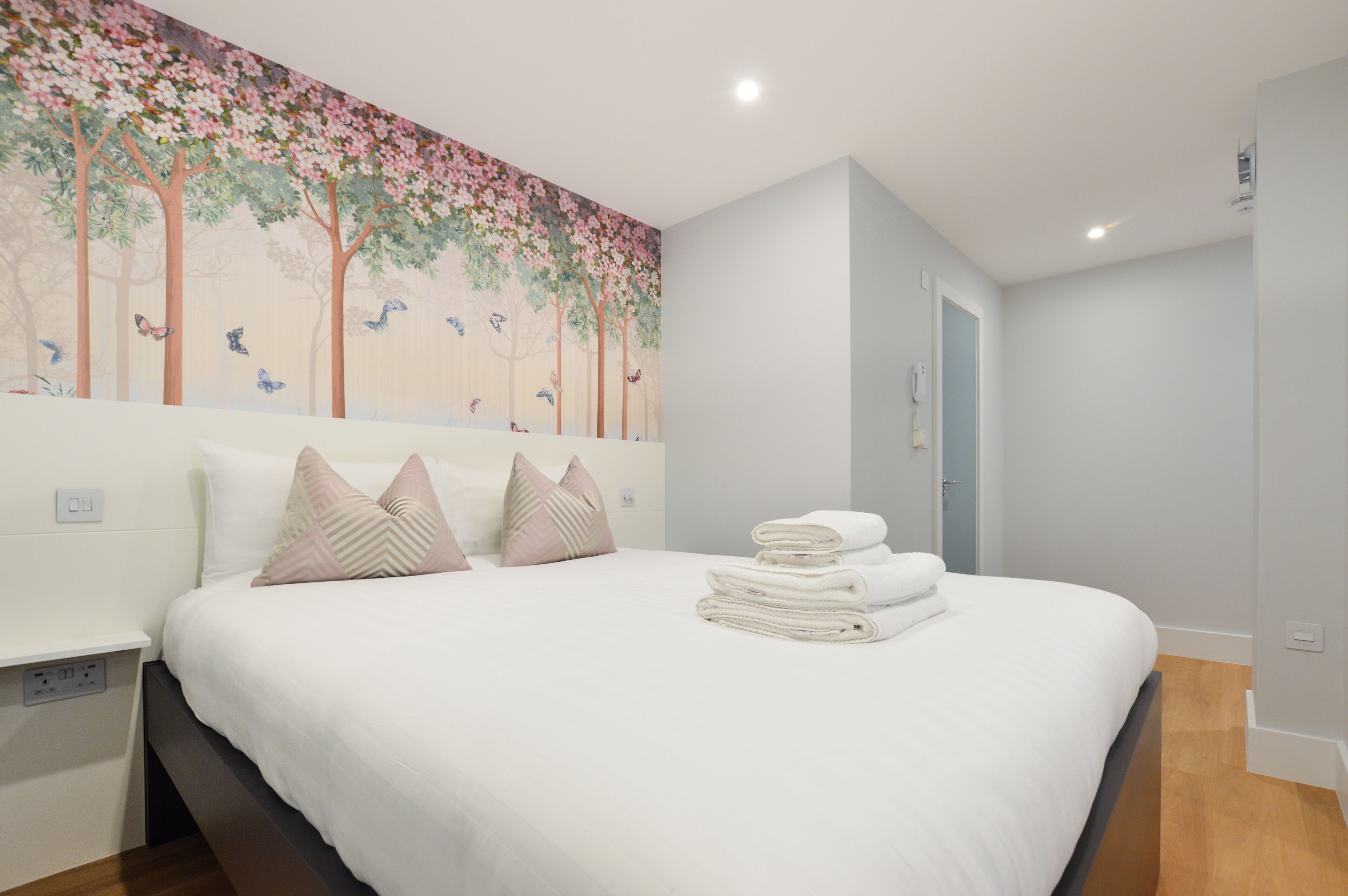 Una cama bien hecha con almohadas y toallas dobladas, un mural de árboles y mariposas, y una habitación de tonos claros con suelo de madera.