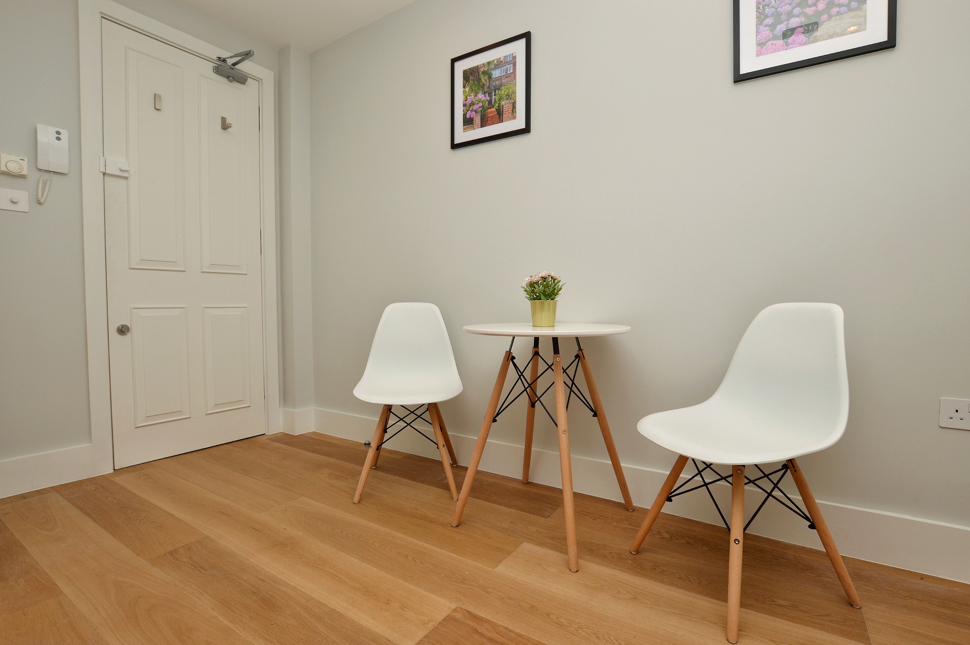 Une pièce avec un plancher en bois, deux chaises blanches, une petite table ronde ornée d’un pot de plante et une porte blanche avec une serrure.