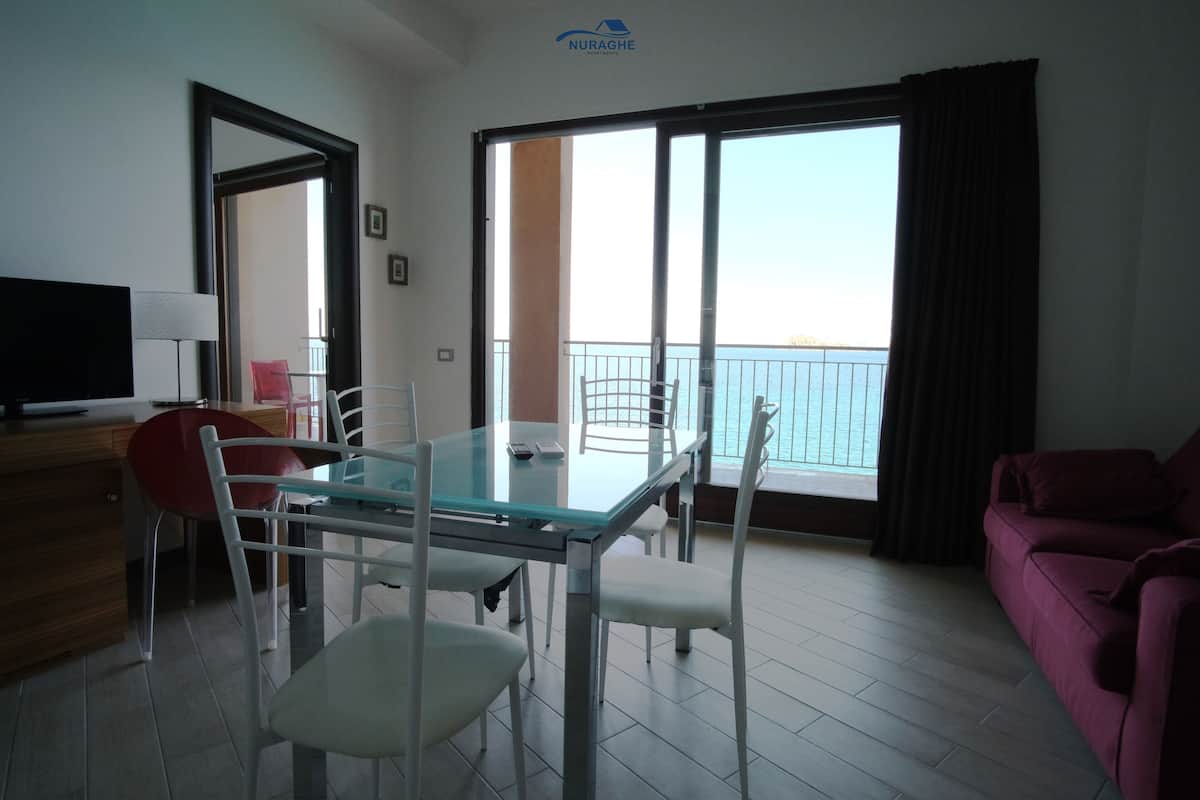A modern living room with a glass dining table, white chairs, a red chair, a pink sofa, a TV, and a balcony with a view.