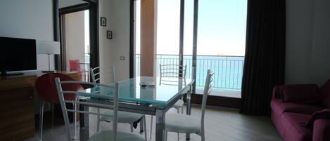 Una sala de estar moderna con una mesa de comedor de vidrio, sillas blancas, una silla roja, un sofá rosa, un televisor y un balcón con vistas.