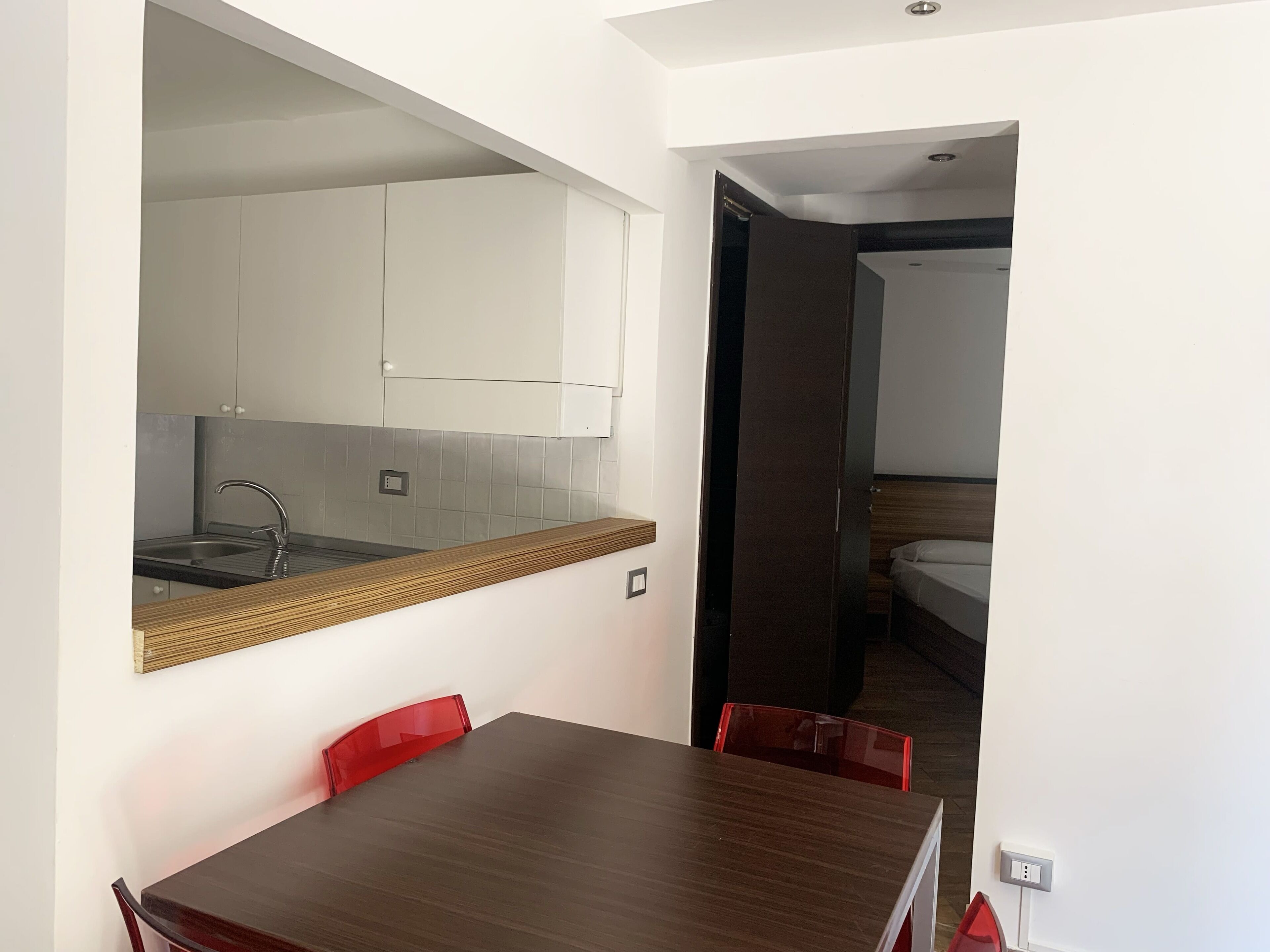 A compact room with a wooden dining table, red chairs, a kitchen area with white cabinets, and a bed in the adjacent room.
