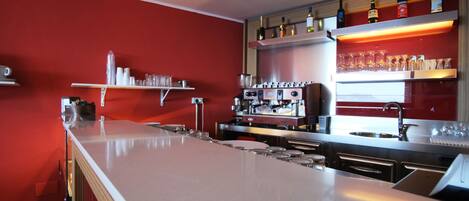 A modern kitchen with a bar area, equipped with a coffee machine, wine bottles, and glasses on shelves.