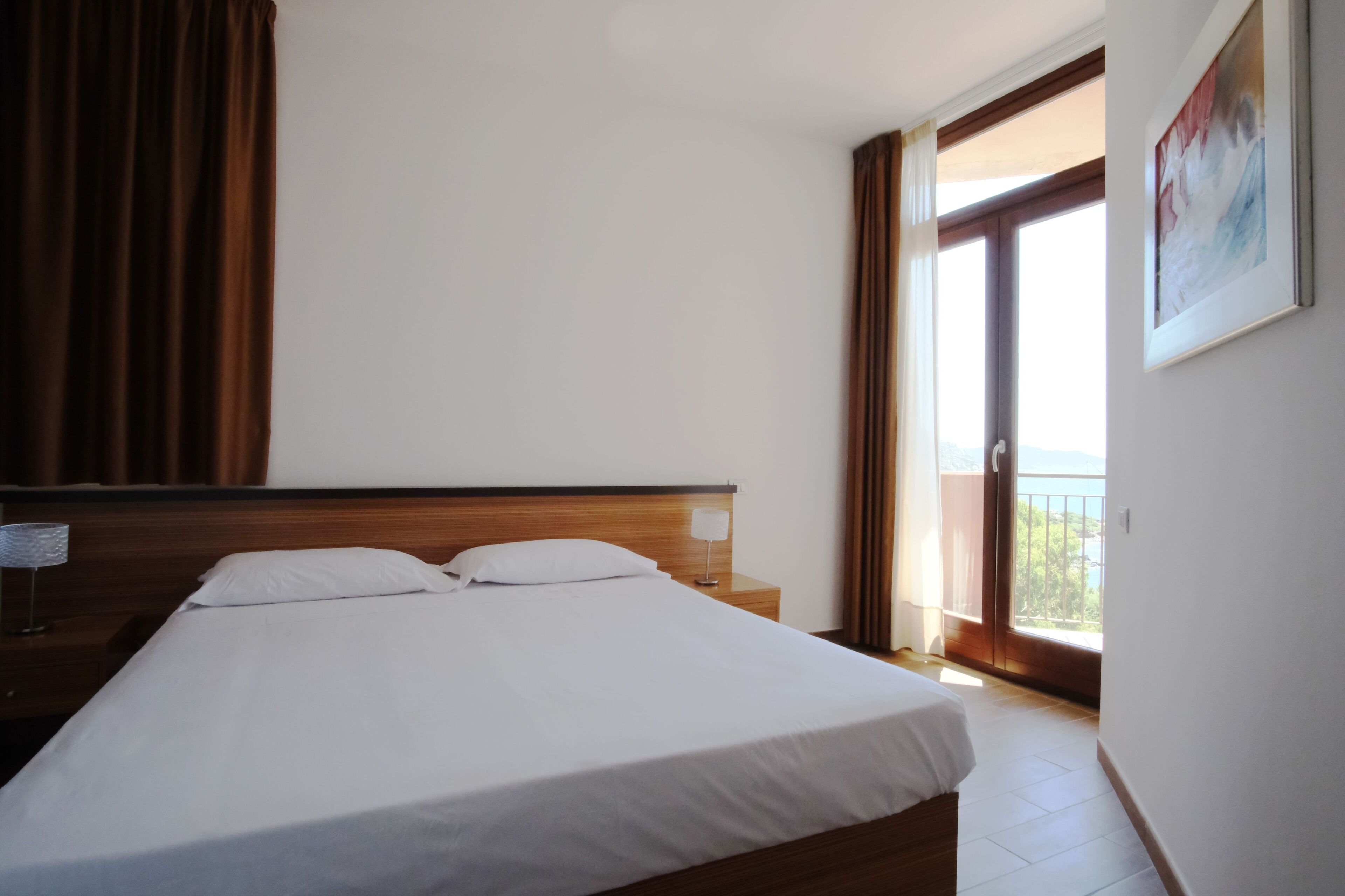 Une chambre d’hôtel avec un grand lit, une table de chevet avec une lampe, une fenêtre avec des rideaux et un tableau au mur.