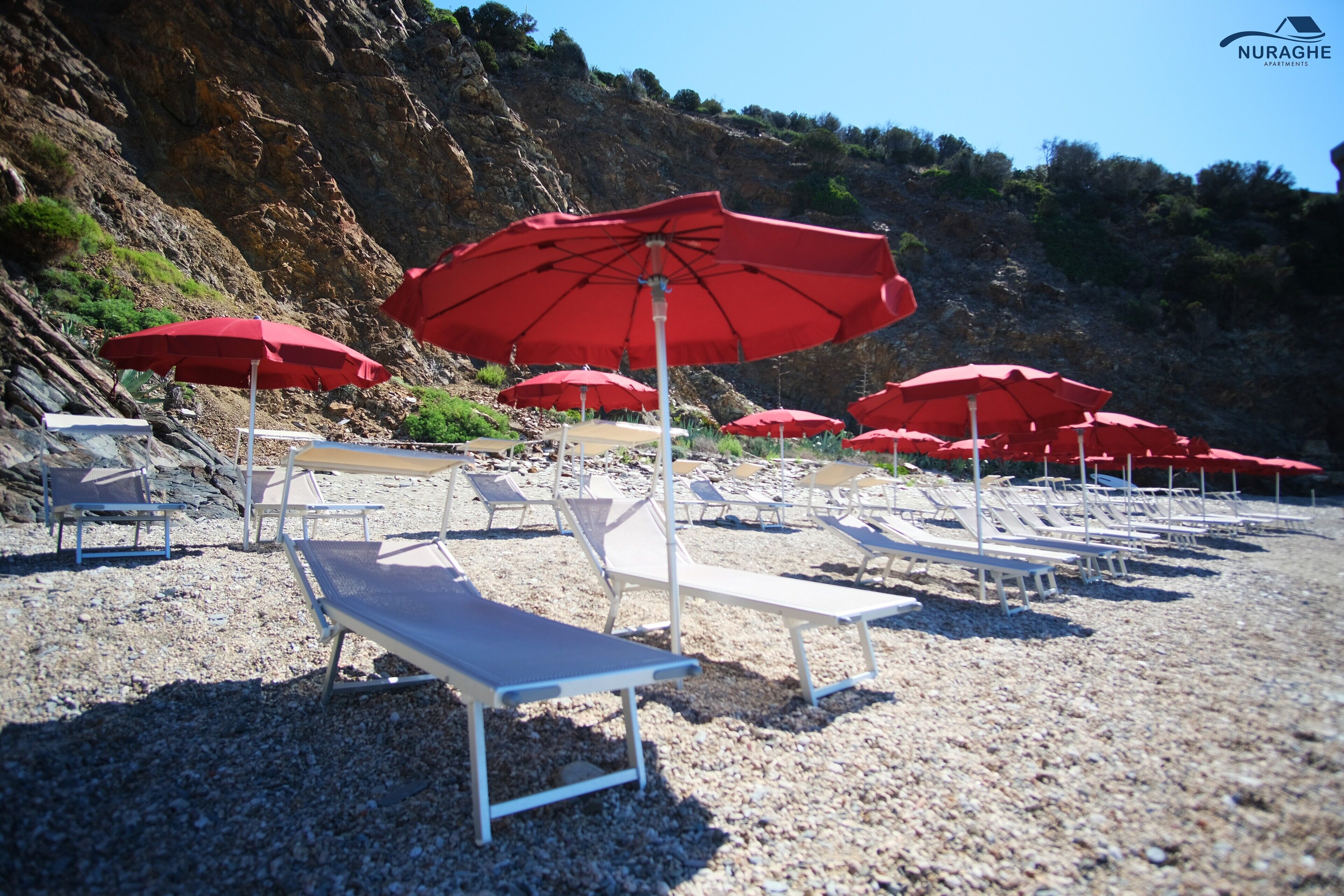 Une plage avec des chaises longues blanches et des parasols rouges, des falaises rocheuses en arrière-plan et un ciel dégagé.