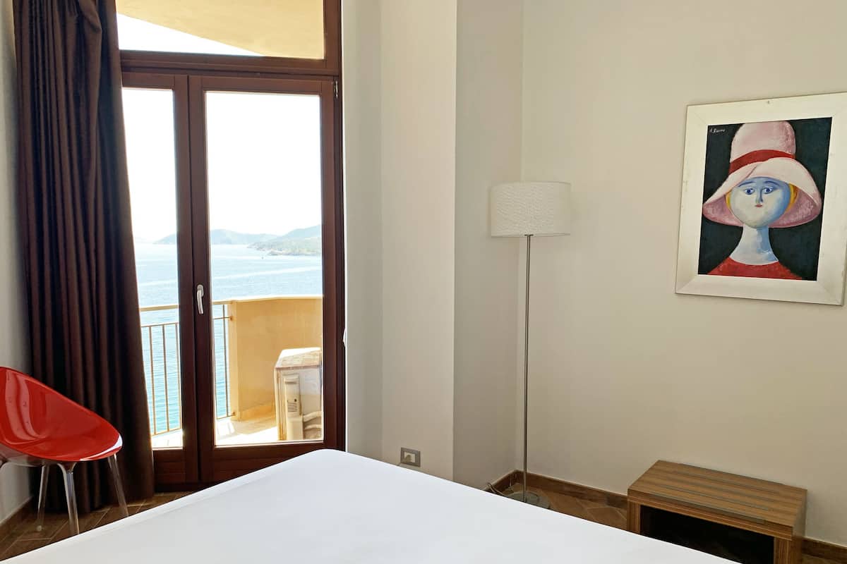 A hotel room with a bed, a red chair, a small wooden bench, a painting on the wall, and a view of the sea through a glass door.
